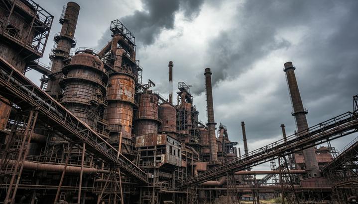 Крупный сталелитейный комбинат с доменными печами, сложной системой труб и дымящими заводскими трубами под пасмурным небом.