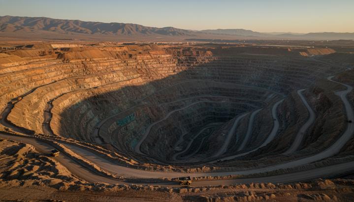 Огромный карьер по добыче меди с террасными уступами, уходящими вглубь земли. По серпантинам едут карьерные самосвалы.